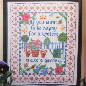 Peut inclure: Broderie au point de croix encadrée dans un cadre noir avec un fond blanc. La broderie représente une scène de jardin avec des fleurs, des papillons et un arrosoir. Le texte dit "Si vous voulez être heureux pour la vie, faites un jardin."