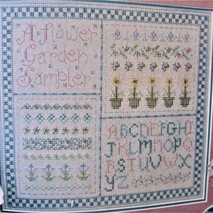 Peut inclure: Un sampler de point de croix encadré avec une bordure à carreaux bleus et blancs. Le sampler présente le texte "A Flower Garden Sampler" et comprend une variété de motifs floraux, dont des tournesols et une rangée de petites fleurs bleues. Le sampler comprend également l'alphabet en point de croix.