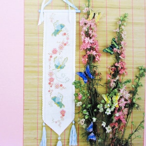 May include: A white cross-stitch wall hanging featuring a blue and green butterfly design. The hanging has blue tassels and is displayed against a bamboo background with pink flowers and blue butterflies.