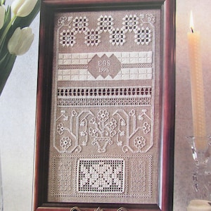 May include: A framed embroidery piece with a white linen background and intricate white stitching. The embroidery features a floral design, a date, and the initials "E.B.S."