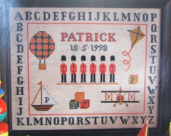 Puede incluir: Bordado de punto de cruz con un globo aerostático rojo, negro y blanco, un velero, un biplano y una fila de cinco guardias de color rojo. El bordado también incluye el nombre "PATRICK" y la fecha "18-5-1998". El alfabeto está bordado alrededor del borde.