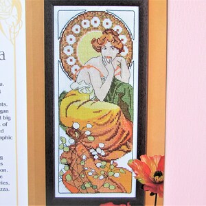 May include: Cross-stitch art featuring a woman in a flowing yellow and orange dress, framed in dark wood. The artwork has a circular design behind the woman's head and is reminiscent of Art Nouveau style.