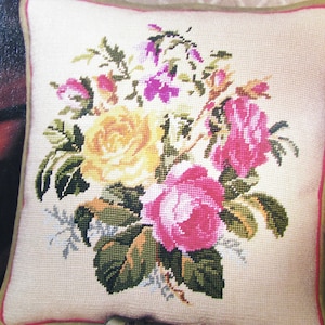 Puede incluir: Una almohada de color crema con un diseño de bordado floral. El bordado presenta rosas rosas, amarillas y moradas con hojas verdes.