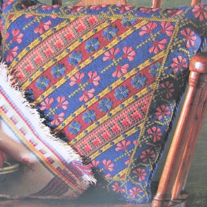 Peut inclure: Un coussin bleu, rouge, vert et jaune à motifs avec une bordure à franges posé sur une chaise en bois.