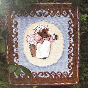 May include: A square, cross-stitched wall hanging featuring a hedgehog holding a cup. The hedgehog is surrounded by a light blue circle and a brown border with floral accents. The background is green.
