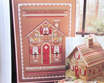 May include: A cross-stitch pattern and a finished decorative house. The pattern features a brown house with red accents, a white lace border, and the text "COTTAGE SWEET COTTAGE." The finished house is a 3D version of the pattern.