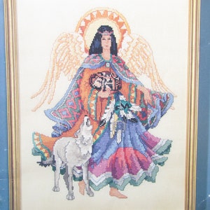 May include: Cross-stitch pattern depicting a Native American angel with a wolf, wearing a colourful dress and holding a drum. The angel has long black hair and a halo. The background is white.