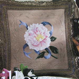 May include: A beige pillow with a brown embroidered frame and a pink flower with blue ribbon design.