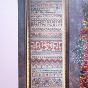 Può includere: Campione di punto croce incorniciato con una cornice color oro. Il campione presenta vari motivi ricamati, tra cui lettere, numeri e disegni floreali in rosso, blu e beige. Il testo include l'alfabeto e i numeri.