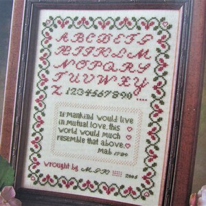 May include: A framed cross-stitch sampler with a white background, green and red embroidery, and the alphabet, numbers, and a quote about love. The quote reads: "If mankind would live in mutual love, this world would much resemble that above." The sampler is signed "wrought by M.FR 2005".