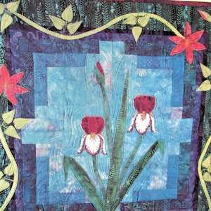 May include: A quilt with a blue background featuring three white and red irises with green leaves. The quilt is framed with a dark green and purple border with a yellow vine and green leaves.