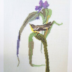 May include: Cross-stitch embroidery of a yellow bird perched on a green branch with purple flowers. The bird is surrounded by green leaves and a brown branch.