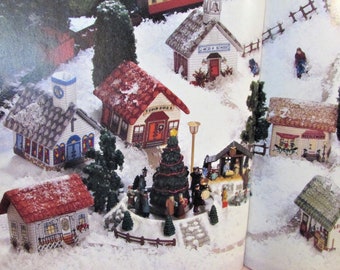 May include: A detailed miniature Christmas village scene with snow-covered buildings, including a church and houses with red roofs. Figures of people are gathered around a Christmas tree and a nativity scene, creating a festive atmosphere.