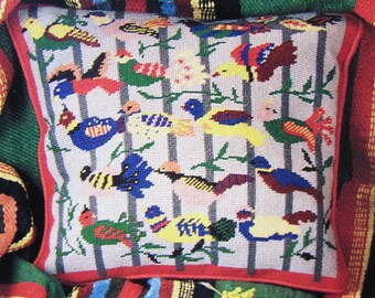 May include: A white pillow with a red border features a colorful needlepoint design of birds on branches. The birds are in shades of blue, red, yellow, green, and black.