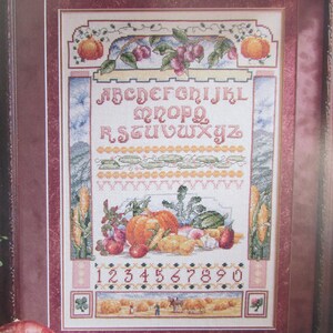 May include: A cross-stitch pattern featuring a colourful alphabet, numbers, and an autumn harvest scene with pumpkins, corn, and other vegetables. The pattern is framed in a brown wood frame.
