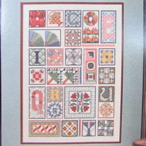 May include: A framed cross-stitch design featuring a patchwork quilt pattern with letters spelling out the word "Welcome". The quilt blocks are in various colours, including red, green, blue, yellow, and pink.