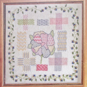 Könnte beinhalten: Ein gerahmtes Kreuzstich-Exemplar mit einer zentralen Blume mit rosa, blauen und grünen Blütenblättern. Das Design umfasst verschiedene geometrische Muster in Pastellfarben, umgeben von einem floralen Rand aus grünen Ranken und blauen Blumen. Der Rahmen ist aus Holz.