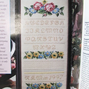 Puede incluir: Un sampler de punto de cruz blanco con un alfabeto enmarcado, diseños florales y la fecha "1997". El sampler incluye instrucciones para la tela, el hilo y los suministros.