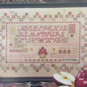 May include: A cross-stitch sampler with a cream-coloured background, featuring a red alphabet, a basket of red berries, and a floral design. The sampler is framed in a dark wood frame. The text "1234567890" is visible at the bottom of the sampler.