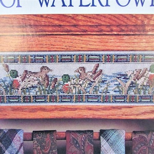 May include: A wooden tie rack with a framed cross-stitch design of ducks and reeds. The cross-stitch is in shades of blue, green, brown, and white. The rack has a brown wooden finish and is holding several ties.