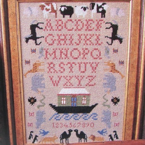 May include: Framed cross-stitch sampler featuring an alphabet, numbers, and various animal and architectural motifs. The design includes cows, sheep, and a depiction of Noah's Ark. The color palette is primarily red, blue, and tan.