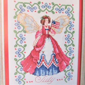 May include: Cross-stitch artwork of a woman with angel wings holding an American flag. The woman is wearing a red and white dress. The word "July" is stitched at the bottom. The artwork is framed with a white and red border.