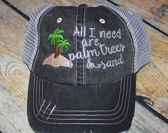 May include: A black and gray trucker hat with a white embroidered design that says "All I need are palm trees & sand". The design also includes two green palm trees on a small island.