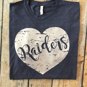 May include: A navy blue t-shirt with a white heart design. The word "Raiders" is printed in a cursive font inside the heart. The shirt is folded on a wooden surface.