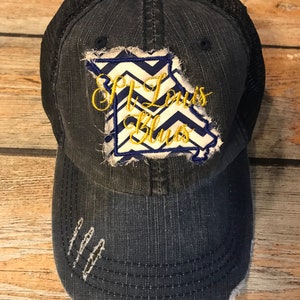 May include: A blue denim trucker hat with a distressed look. The hat features a black mesh back and a patch with a white and blue chevron pattern and the text "St. Louis Blues" in gold embroidery.