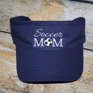 May include: A navy blue visor with white embroidery that reads "Soccer Mom" and a white soccer ball graphic.