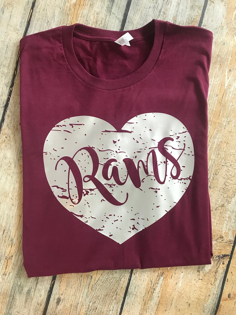 May include: A maroon t-shirt with a distressed silver heart design. The word "Rams" is printed in a cursive font within the heart. The shirt is folded and lies on a wooden surface.