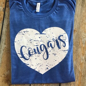 May include: A blue t-shirt with a white heart-shaped graphic. The word "Cougars" is written in a script font inside the heart. The shirt is folded and has a tag that says "Canvas."