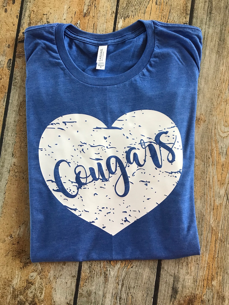May include: A blue t-shirt with a white heart-shaped graphic. The word "Cougars" is written in a script font inside the heart. The shirt is folded and has a tag that says "Canvas."