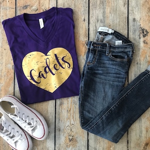 May include: Purple V-neck t-shirt with a gold heart that says "Cadets" on it. The shirt is paired with blue jeans and white sneakers.