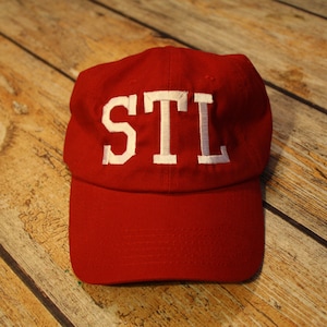 Puede incluir: Una gorra de béisbol roja con un "STL" bordado en blanco en la parte delantera.