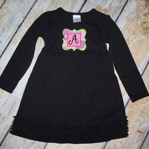 Könnte beinhalten: Schwarzes Langarmkleid mit Rüschen am Saum. Das Kleid hat ein buntes Stickmuster mit dem Buchstaben "A" in der Mitte.