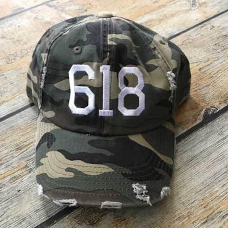 May include: Camouflage baseball cap with white distressed lettering that reads "618".
