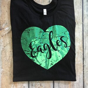 May include: Black t-shirt with a green heart-shaped design. The word "Eagles" is written in a cursive font within the heart. The shirt is folded on a wooden surface.