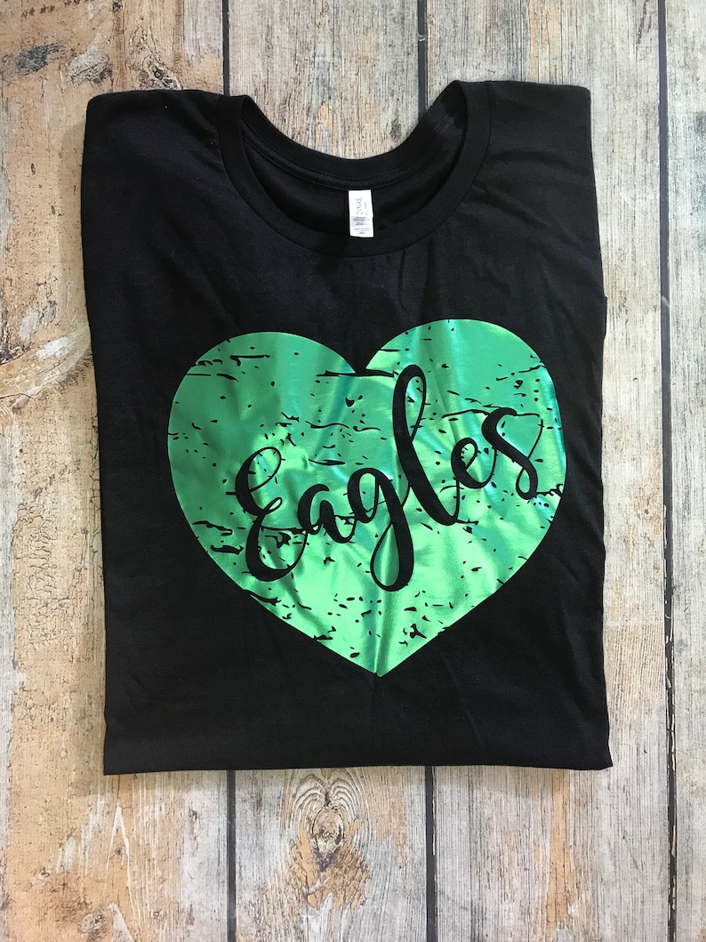 May include: Black t-shirt with a green heart-shaped design. The word "Eagles" is written in a cursive font within the heart. The shirt is folded on a wooden surface.