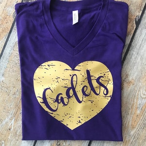 May include: A purple V-neck t-shirt with a gold heart design. The heart has a distressed texture and the word "Cadets" in a script font. The shirt is folded neatly on a wooden surface.