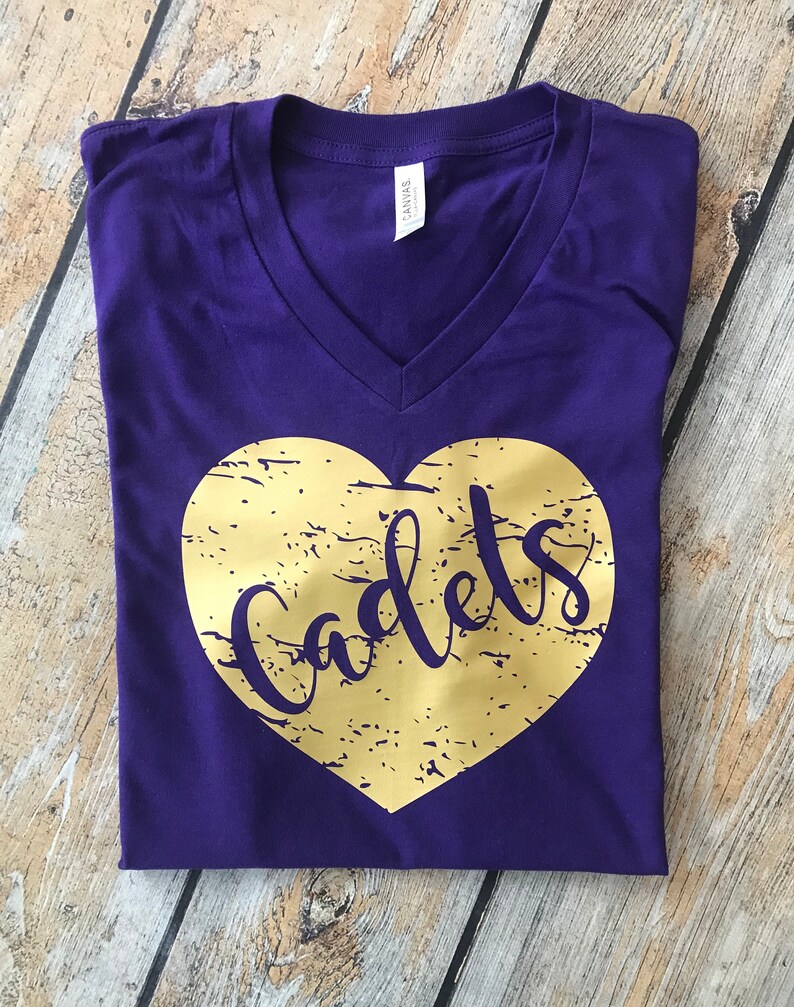 May include: A purple V-neck t-shirt with a gold heart design. The heart has a distressed texture and the word "Cadets" in a script font. The shirt is folded neatly on a wooden surface.