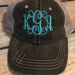 May include: A black and gray distressed trucker hat with a gray mesh back. The hat has a turquoise monogram of the letters "KGS" embroidered on the front.