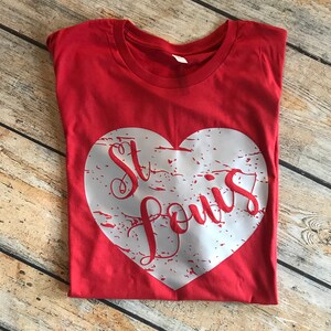 May include: A red t-shirt with a white heart-shaped graphic. The graphic features the words "St. Louis" in a red, cursive font. The shirt is folded and lies on a wooden surface.