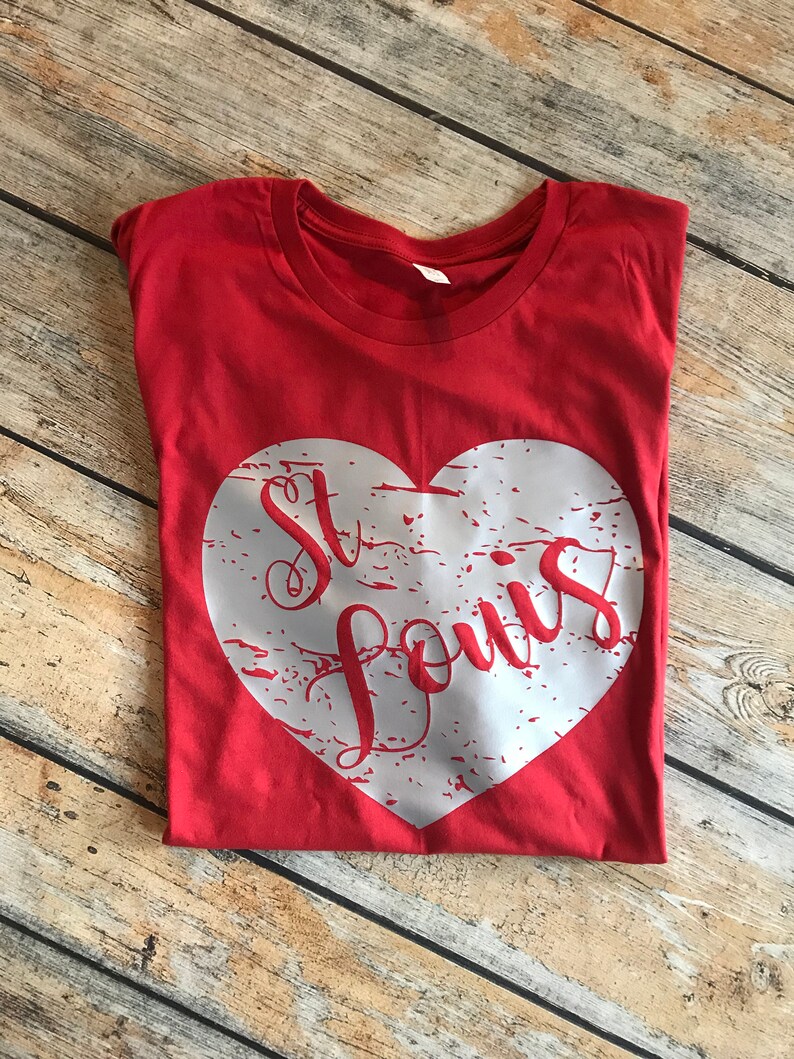 May include: A red t-shirt with a white heart-shaped graphic. The graphic features the words "St. Louis" in a red, cursive font. The shirt is folded and lies on a wooden surface.