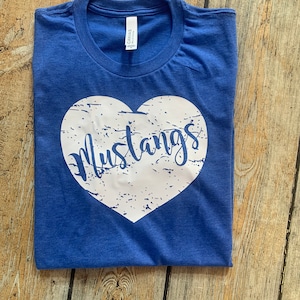 May include: Royal blue t-shirt with a white heart-shaped graphic. The heart contains the word "Mustangs" in a distressed, cursive font. The shirt is folded neatly on a wooden surface.