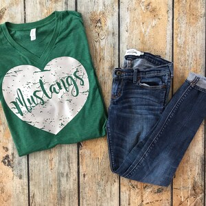 May include: A green V-neck t-shirt with a white heart design that says "Mustangs" and a pair of blue denim jeans. The jeans are folded at the ankles. The items are on a wooden surface.