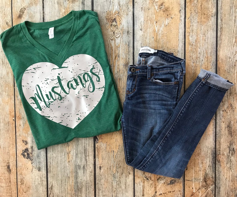 May include: A green V-neck t-shirt with a white heart design that says "Mustangs" and a pair of blue denim jeans. The jeans are folded at the ankles. The items are on a wooden surface.