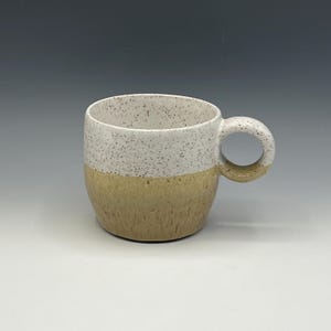 以下が含まれることがあります： 白の斑点模様の上部とタン色の下部が特徴的な、手作りのセラミックマグカップ。丸みを帯びたハンドルとシンプルでモダンなデザインです。コーヒー、紅茶、その他の飲み物に最適です。