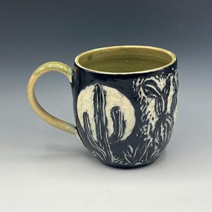 May include: A ceramic mug with a dark navy blue glaze featuring a carved cream-colored desert scene with a large moon and saguaro cacti. The interior and the handle are finished in a smooth, light olive green glaze, providing a contrast.