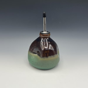 May include: A handcrafted ceramic oil bottle with a stainless steel spout and a black stopper. The bottle features a brown top, a light green base, and a tan band in between. The bottle is designed for dispensing oil.